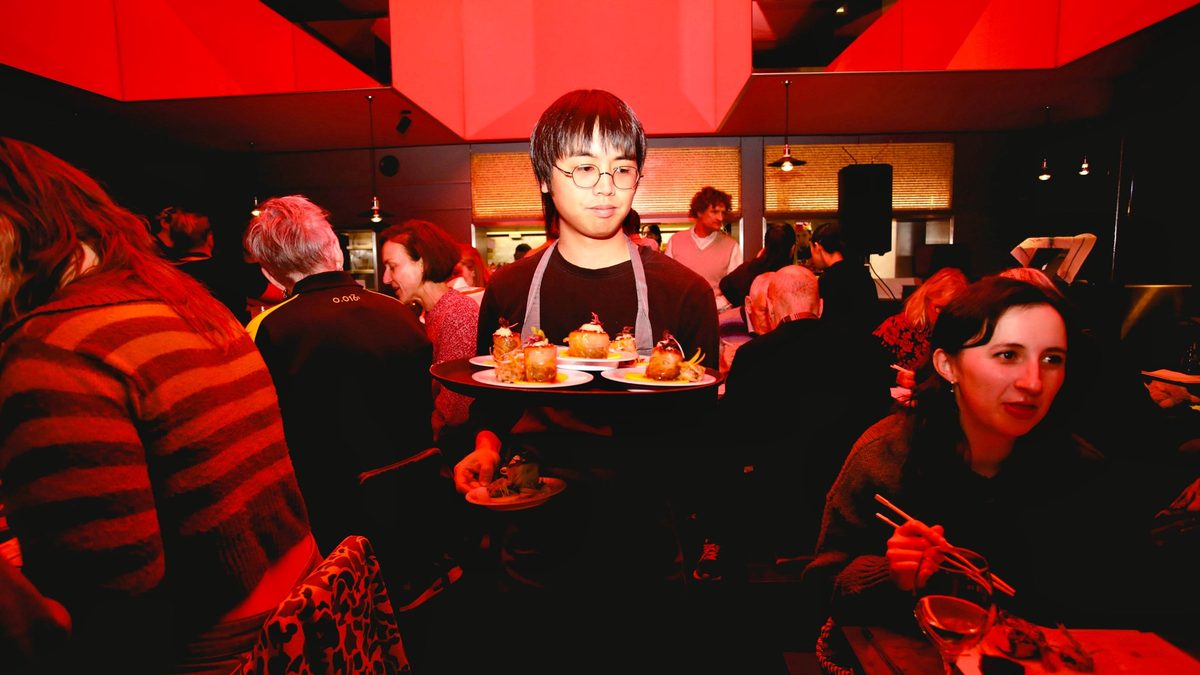 A server carries a tray of plates through a packed event under the red ceiling