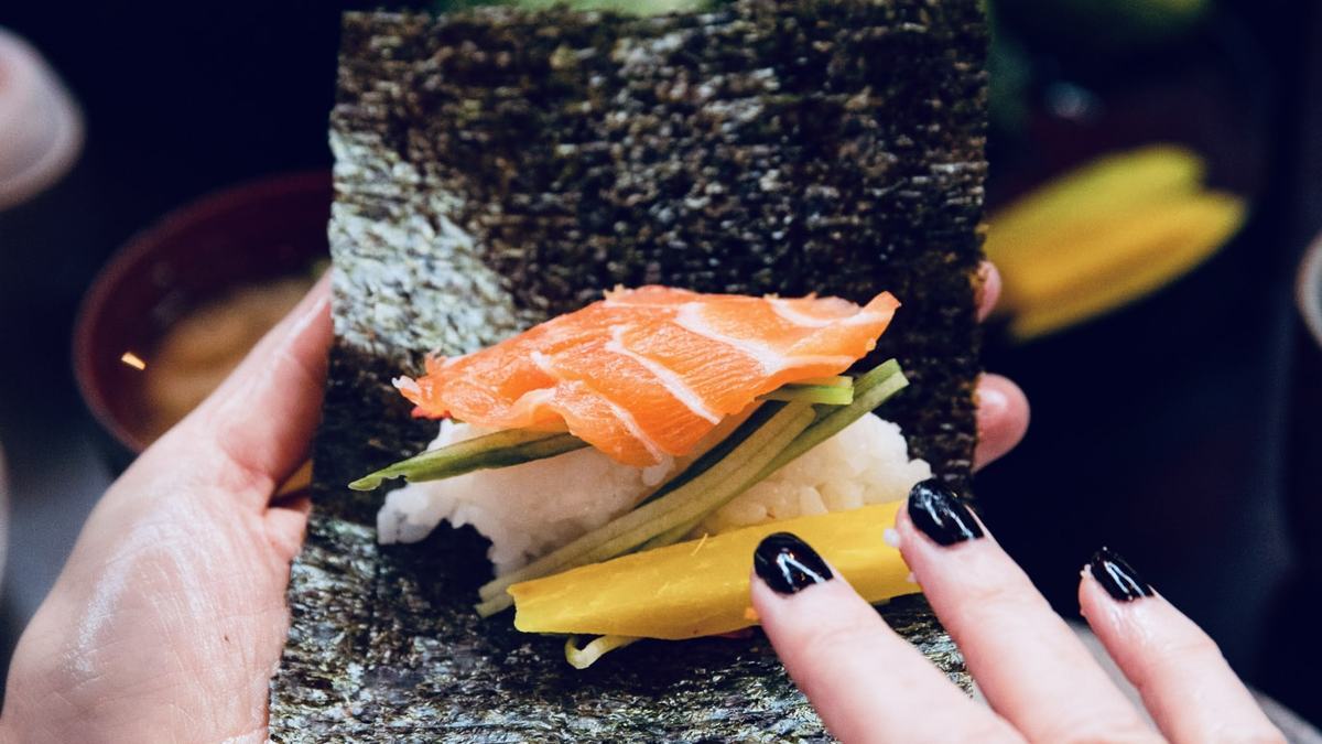 Hands building a temaki hand roll with nori, rice, salmon and cucumber